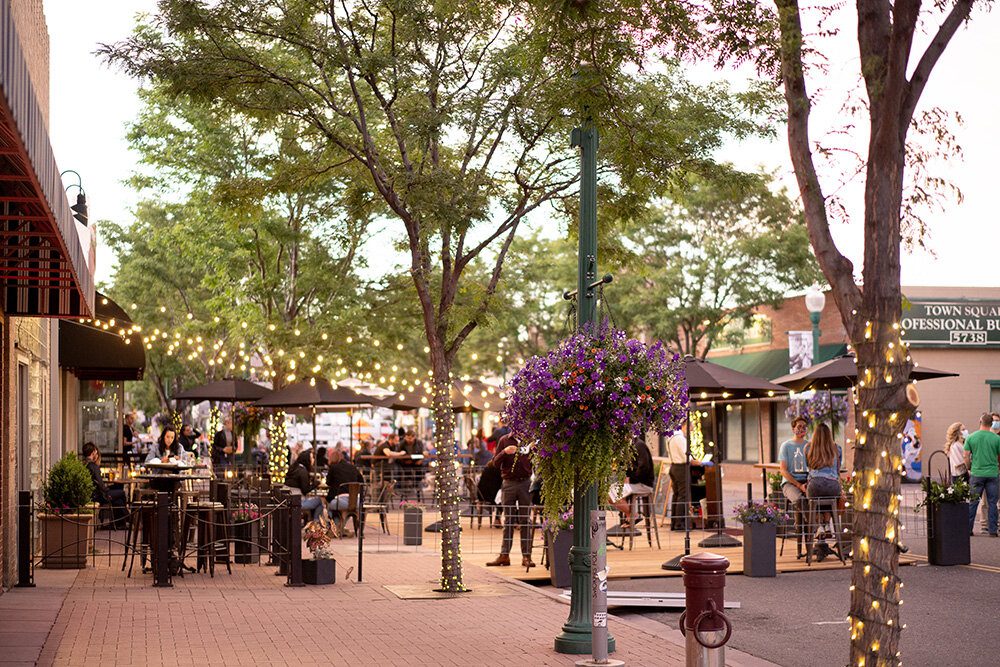 Evening view of a vibrant downtown area in Arvada, Colorado, decorated with string lights and bustling with people at outdoor cafes and restaurants.