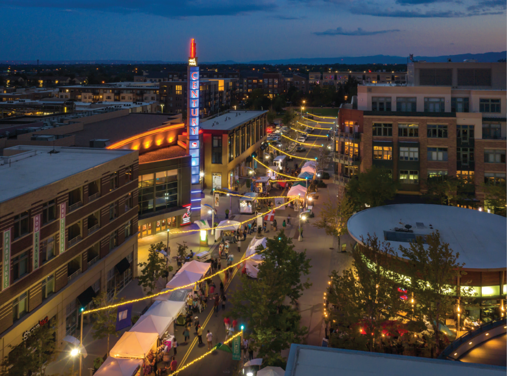 Aerial view of Belmar shopping district in Lakewood, Colorado, during an evening market event with illuminated string lights and people walking between stalls.