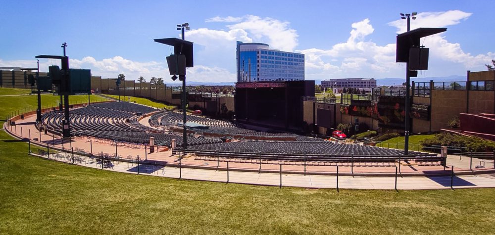 A spacious outdoor concert venue with rows of seats, a stage, and a modern building in the background under a clear blue sky.